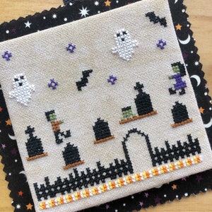 Op de afbeelding: Een wit borduurwerkontwerp op een beige stof met een zwart-wit Halloween-thema. Het ontwerp toont een begraafplaats met grafstenen, een hek, een poort, geesten, vleermuizen en een heks. De stof is omlijst door een zwarte stof met witte sterren.
