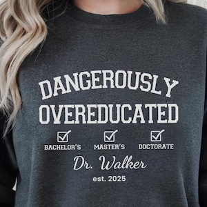 May include: Dark grey heather sweatshirt with the text "DANGEROUSLY OVEREDUCATED" in white. The design includes checkmarks for Bachelor's, Master's, and Doctorate degrees, along with "Dr. Walker est. 2025". A "Personalized ITEM" stamp is in the upper right corner.