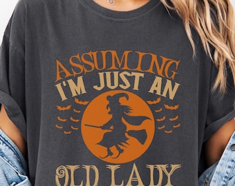 Puede incluir: Camiseta gris oscuro con un diseño de Halloween. El diseño presenta una bruja en una escoba frente a una luna naranja, rodeada de murciélagos, y el texto "Assuming I'm Just An Old Lady Was Your First Mistake."