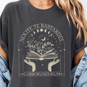May include: Dark grey t-shirt with a beige graphic of an open book with plant life growing from it, held by hands. The text "NOLITE TE BASTARDES" arches above the book, and "CARBORUNDORUM" is below.