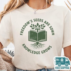 Puede incluir: Camiseta color crema con un gráfico verde de un libro abierto con un árbol que crece de él. El texto alrededor del gráfico dice "FREEDOM'S SEEDS ARE SOWN" y "KNOWLEDGE GROWS". La camiseta es de un material suave.