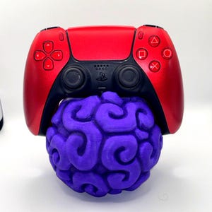 May include: A red and black video game controller rests on top of a purple, brain-shaped object. The controller has the standard button layout with a PlayStation logo. The brain-shaped object has a detailed, textured surface.
