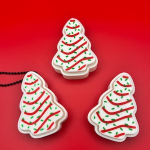 May include: Three Christmas tree-shaped ornaments with white bases, red stripes, and green accents. The ornaments are on a red background. A black beaded necklace is partially visible in the image.