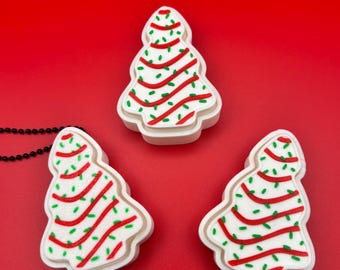 May include: Three Christmas tree-shaped ornaments with white bases, red stripes, and green accents. The ornaments are on a red background. A black beaded necklace is partially visible in the image.