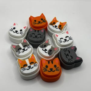 May include: A collection of nine colorful cat-shaped eraser toppers. The toppers are made of foam and feature different colored cats with smiling faces. The cats are in various colors, including white, orange, gray, and black.