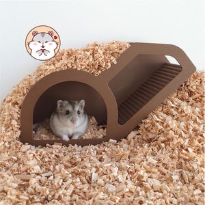 May include: A small gray and white hamster sits inside a brown plastic tunnel hideout with a ramped roof. The structure is surrounded by light brown wood shavings. A circular graphic of a cartoon hamster with a heart is visible in the top left.