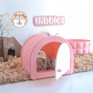 May include: A light pink arched hamster house with a white door, featuring two small hamsters inside. The structure has a ribbed roof design. A sign above reads "Nibbles" in white letters on a pink background, accompanied by a circular hamster icon.
