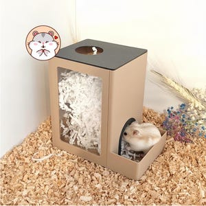 Puede incluir: Un escondite rectangular beige para hámster con techo gris oscuro, una abertura circular superior y una ventana frontal transparente llena de lecho de papel blanco. Un pequeño hámster claro entra por la entrada lateral arqueada.