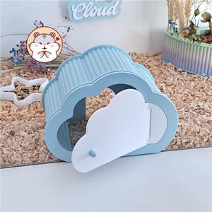 May include: A light blue cloud-shaped hamster house with a white door featuring a small blue knob. The structure has a ribbed roof and a hollow interior. A circular sticker with a cartoon hamster and the word Cloud are visible in the background.