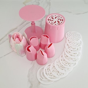 Puede incluir: Un conjunto de herramientas rosas para decorar pasteles, que incluye boquillas, un molde con forma de flor, un soporte pequeño y varias plantillas blancas con diseños. Las herramientas están dispuestas sobre una superficie blanca, lo que sugiere un tema de repostería o artesanía.