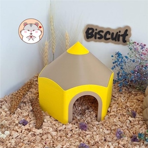 May include: A small animal hideout shaped like a yellow and tan pencil with a pointed top and an arched entrance. The structure is placed on wood shavings and features a circular sticker of a hamster and a sign with the text Biscuit.