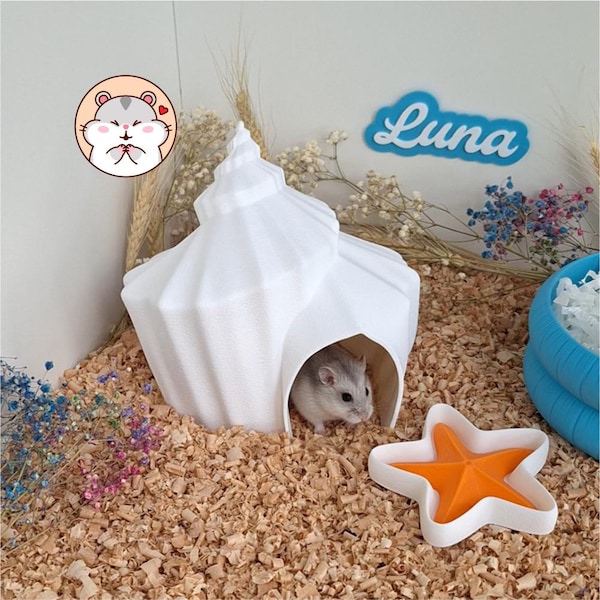 May include: A white seashell-shaped hamster hideout with a small opening, featuring a grey and white hamster inside. Next to it is an orange and white starfish-shaped dish. A blue sign with the text Luna is visible in the background.