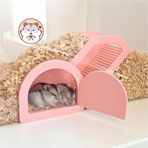 May include: A pink plastic hamster house with a clear front panel and an open side door, partially buried in light brown wood shavings. Two small grey and white hamsters are sleeping inside the structure. A circular icon of a hamster is visible.