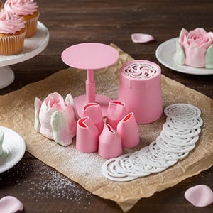 Op de afbeelding: Een roze taartdecoratieset met verschillende spuitmonden, een standaard en sjablonen. De set is gerangschikt op een bruin oppervlak met cupcakes en bloemvormige lekkernijen.