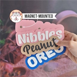 Puede incluir: Un conjunto de letreros superpuestos con bordes festoneados y soportes magnéticos. La capa superior rosa muestra un icono de hámster y el texto MAGNET-MOUNTED. Debajo hay letras blancas que forman Nibbles, una capa beige con texto en cursiva negra Peanut y una capa azul con letras blancas OREO.