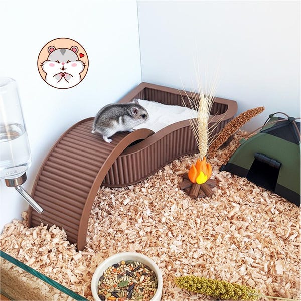 May include: A small gray and white hamster stands on a brown, ribbed, arched bridge over a white sand bath. Nearby is a miniature green tent, a small decorative campfire with a wheat stalk, a water bottle, and a bowl of mixed seeds.