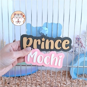 May include: A custom name sign for a pet cage featuring the text "prince" in gold on a dark grey scalloped background and "Mochi" in white on a pink scalloped background, attached to a vertical metal wire cage with a blue plastic fastener.