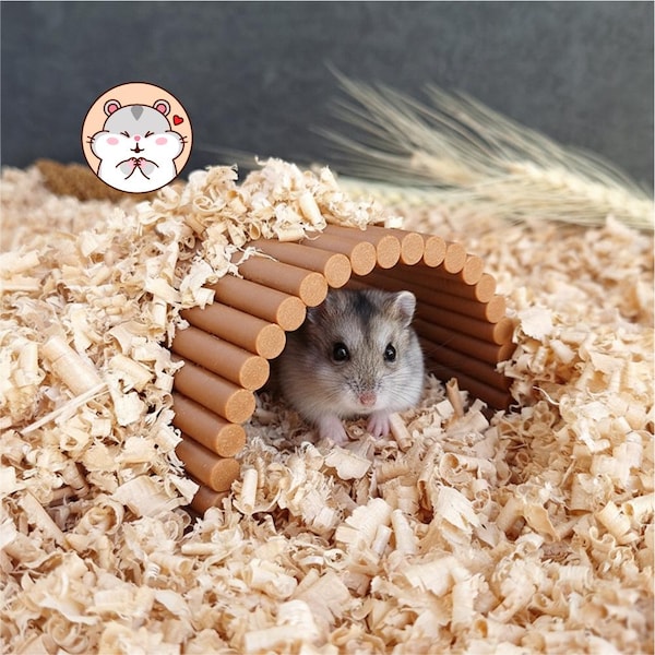 May include: A small gray and white hamster sits inside a brown, arched wooden tunnel made of connected cylindrical logs. The tunnel is partially covered in light-colored wood shavings, and a cartoon hamster icon is visible in the corner.