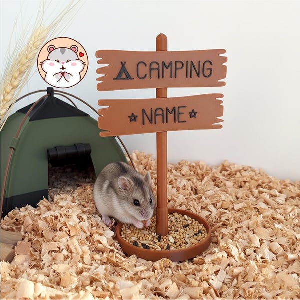 May include: A brown plastic signpost with two horizontal boards featuring the text CAMPING and NAME, standing in a circular brown base filled with seeds. A small hamster sits next to the base, near a green tent-shaped structure.