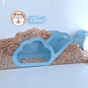 May include: A light blue cloud-shaped hamster hideout with a ramp and a cutout design, partially buried in wood shavings. A circular sticker shows a cartoon hamster, and a blue sign with the white text Cloud is mounted on the clear wall.