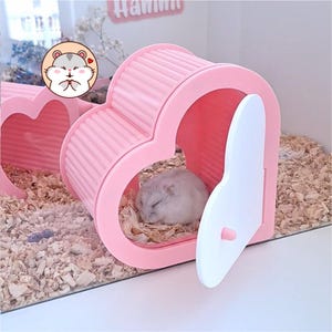 May include: A light pink heart-shaped hamster house with a white door and a ribbed roof. A small grey hamster is sleeping inside the structure, which is placed on a bed of wood shavings. A circular sticker with a cartoon hamster is visible.