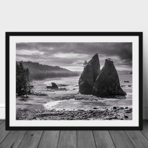 May include: Black and white photograph of two large rock formations on a beach. The rocks are tall and narrow, and they are separated by a small gap. The beach is covered in sand and rocks, and the water is calm and still. The sky is cloudy and overcast.