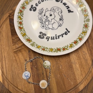 Puede incluir: Un plato blanco con un gráfico de ardilla y el texto "Second-Hand Squirrel" descansa sobre una tabla de cortar de madera. En primer plano, una pulsera plateada con tres colgantes redondos estampados. Los colgantes presentan diseños florales azules, verdes y amarillos.