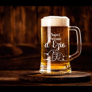 Peut inclure: Une chope à bière en verre transparent avec une poignée. La chope est remplie d'une bière jaune dorée. Le texte "Super Parrain d'Ezio" est gravé sur le devant de la chope.