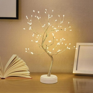 May include: A decorative LED tree lamp with warm white lights. The tree has a silver trunk and branches, with small lights at the tips. It sits on a white circular base. An open book and a picture frame are in the background.