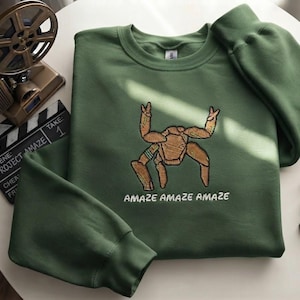 May include: An olive green crewneck sweatshirt featuring an embroidered tan, multi-legged creature with raised arms. Below the creature, the text "AMAZE AMAZE AMAZE" is embroidered in white thread. The garment has long sleeves and a ribbed collar.