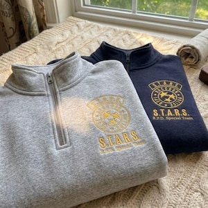 May include: A light gray and a dark navy blue quarter-zip sweatshirt. Each sweatshirt features an embroidered gold circular emblem with the text S.T.A.R.S. Raccoon Police Dept and the words S.T.A.R.S. R.P.D. Special Team printed below the logo.