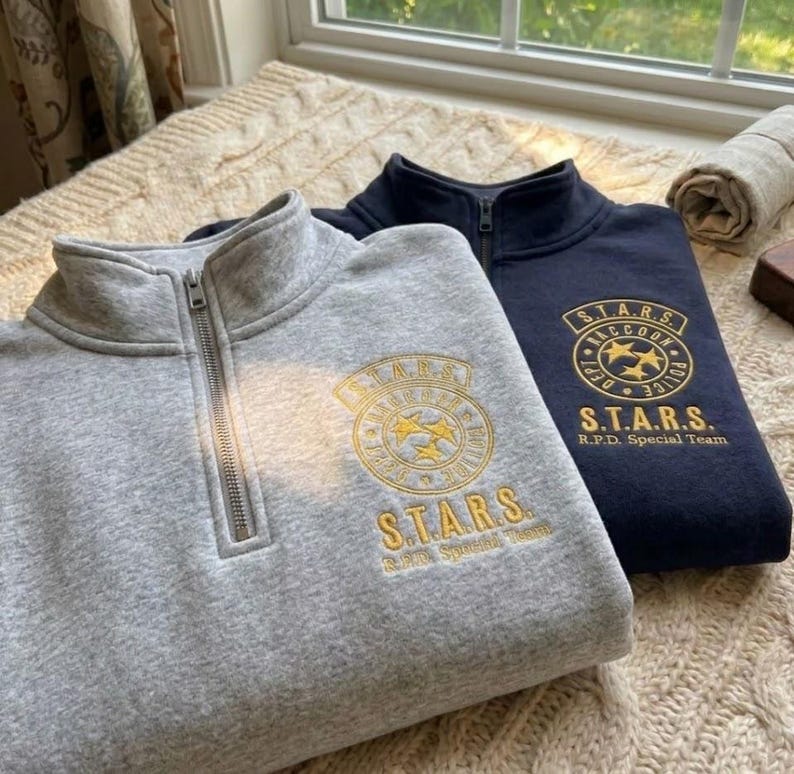 May include: A light gray and a dark navy blue quarter-zip sweatshirt. Each sweatshirt features an embroidered gold circular emblem with the text S.T.A.R.S. Raccoon Police Dept and the words S.T.A.R.S. R.P.D. Special Team printed below the logo.
