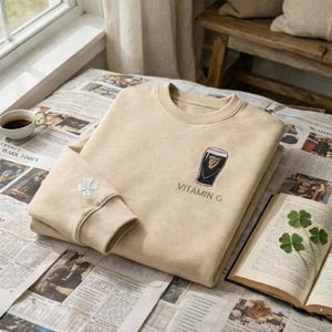 May include: Beige sweatshirt with a crew neck and long sleeves. The front features an embroidered black and gold beer glass design with the text "VITAMIN G". A small embroidered four-leaf clover is on the left sleeve.