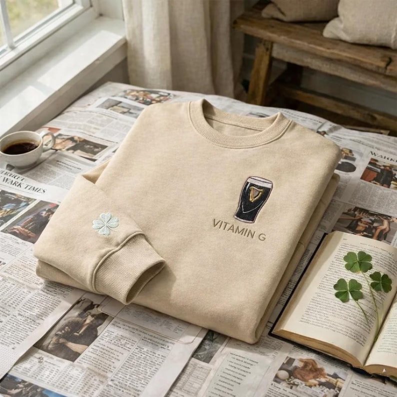 May include: Beige sweatshirt with a crew neck and long sleeves. The front features an embroidered black and gold beer glass design with the text "VITAMIN G". A small embroidered four-leaf clover is on the left sleeve.