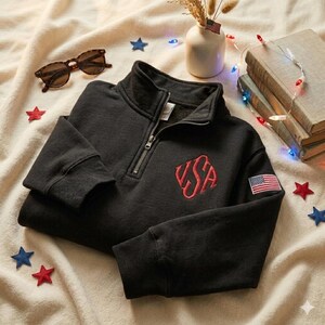 May include: Black quarter-zip sweatshirt with a red embroidered "USA" design on the chest and an American flag patch on the sleeve. The sweatshirt has a ribbed collar and cuffs. Tortoiseshell sunglasses and decorative stars are also in the image.