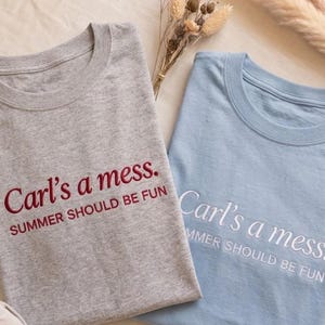 May include: Two t-shirts, one gray and one light blue, are folded and displayed. The gray shirt has the embroidered text "Carl's a mess. SUMMER SHOULD BE FUN" in burgundy. The light blue shirt has the same text embroidered in white.