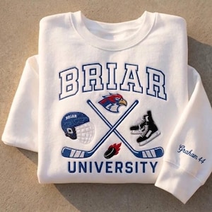 May include: A white crewneck sweatshirt featuring embroidered blue text that reads BRIAR UNIVERSITY. The design includes a blue hockey helmet, a bird mascot head, black ice skates, a hockey puck, and two crossed hockey sticks. The sleeve displays the text Graham 44.