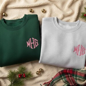 Könnte beinhalten: Zwei Sweatshirts mit Rundhalsausschnitt, eines in Waldgrün und eines in Hellgrau, jeweils mit einem rosa Monogramm bestickt. Die Sweatshirts werden auf einer cremefarbenen, strukturierten Oberfläche mit goldenen Glocken und Grünzweigen präsentiert.