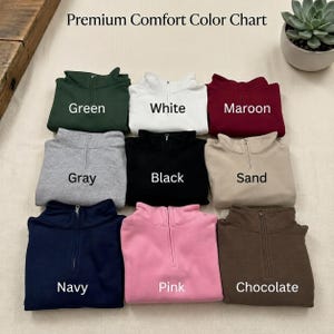 May include: A Premium Comfort Color Chart displaying nine quarter-zip sweatshirts in green, white, maroon, gray, black, sand, navy, pink, and chocolate. Each garment features a central zipper and is labeled with its corresponding color name.