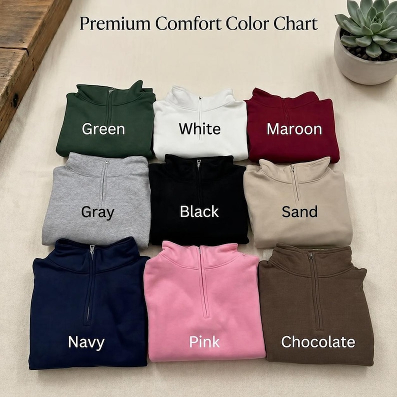May include: A Premium Comfort Color Chart displaying nine quarter-zip sweatshirts in green, white, maroon, gray, black, sand, navy, pink, and chocolate. Each garment features a central zipper and is labeled with its corresponding color name.