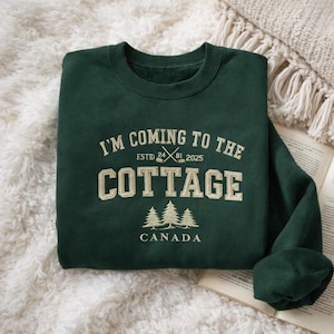 Może przedstawiać: Ciemnozielona bluza z kremowym napisem "I'M COMING TO THE COTTAGE". Poniżej trzy sosny i słowo "CANADA". Bluza jest złożona na białej, puszystej powierzchni, z książką w tle.