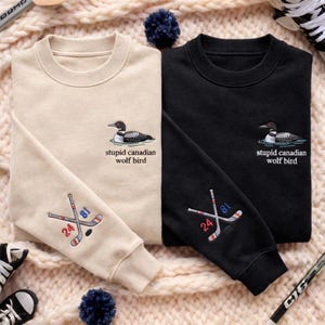 May include: Two crewneck sweatshirts, one beige and one black, with embroidered designs. Each features a loon and the text "stupid canadian wolf bird." The beige sweatshirt has hockey sticks with the numbers 24 and 81 on the sleeve.
