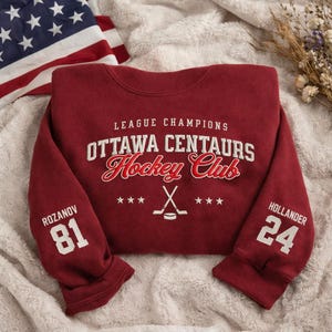 May include: A maroon Ottawa Centaurs Hockey Club sweatshirt with white lettering and graphics. The sleeves display the names and numbers of players Rozanov (81) and Hollander (24). An American flag is visible in the background.