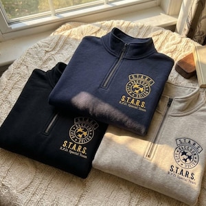 May include: Three quarter-zip sweatshirts in navy blue, black, and light gray. Each features a circular embroidered patch on the chest with the text S.T.A.R.S. Raccoon Police Dept and three stars in the center, with S.T.A.R.S. R.P.D. Special Team below.