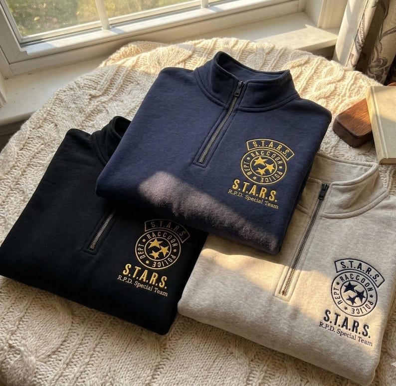 May include: Three quarter-zip sweatshirts in navy blue, black, and light gray. Each features a circular embroidered patch on the chest with the text S.T.A.R.S. Raccoon Police Dept and three stars in the center, with S.T.A.R.S. R.P.D. Special Team below.