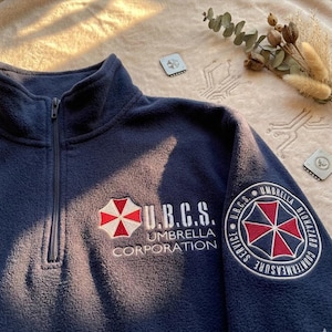 Może przedstawiać: Granatowy polar z zamkiem błyskawicznym do połowy klatki piersiowej, z wyhaftowanym czerwono-białym logo Umbrella Corporation i napisem U.B.C.S. na piersi oraz okrągłą naszywką U.B.C.S. Umbrella Biohazard Countermeasure Service na lewym rękawie.