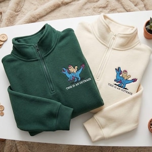 May include: A dark green and a cream-coloured quarter-zip sweatshirt, each featuring an embroidered graphic of a small cat riding a blue dinosaur with the text "THIS IS AN OUTRAGE" printed underneath the design in a clean, black sans-serif font.