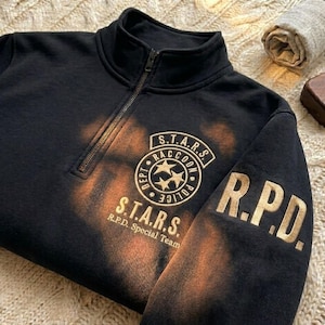 May include: A black quarter-zip sweatshirt featuring a distressed orange bleach-effect pattern. The chest displays a circular S.T.A.R.S. Raccoon Police Dept logo with three stars, and the sleeve features the gold-colored text R.P.D. in block letters.