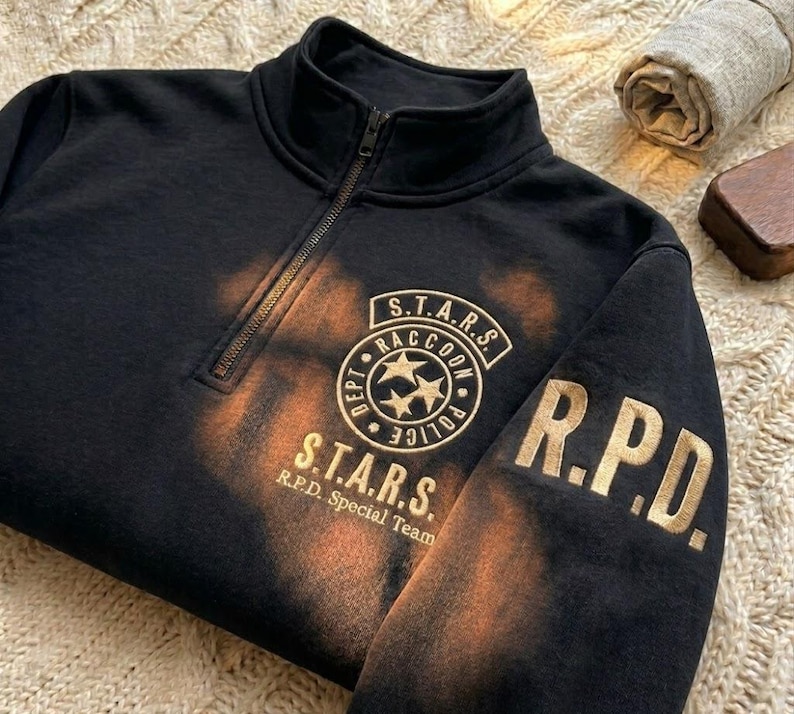 May include: A black quarter-zip sweatshirt featuring a distressed orange bleach-effect pattern. The chest displays a circular S.T.A.R.S. Raccoon Police Dept logo with three stars, and the sleeve features the gold-colored text R.P.D. in block letters.