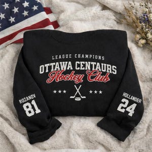Puede incluir: Sudadera negra con texto blanco y rojo "LEAGUE CHAMPIONS OTTAWA CENTAURS Hockey Club". Las mangas muestran los nombres y números "ROZANOV 81" y "HOLLANDER 24". También hay un gráfico de palo y disco de hockey.